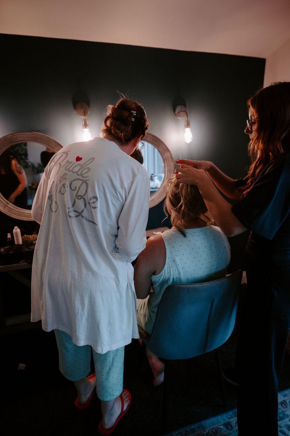 Bride-to-be in white robe gets hair styled by professional stylist in warmly lit bridal preparation room.