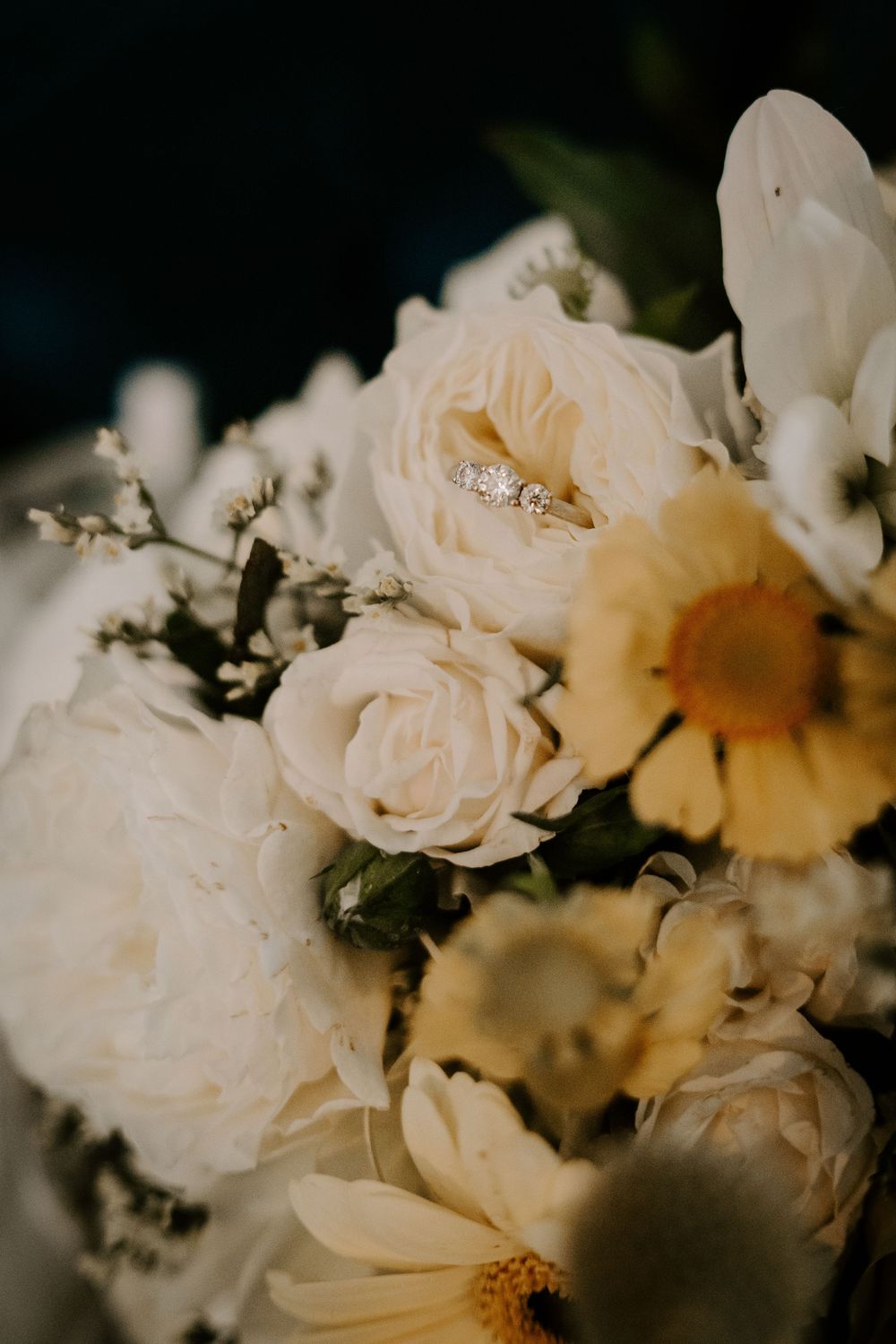 Diamond engagement ring resting on white roses and yellow daisies in bridal bouquet.