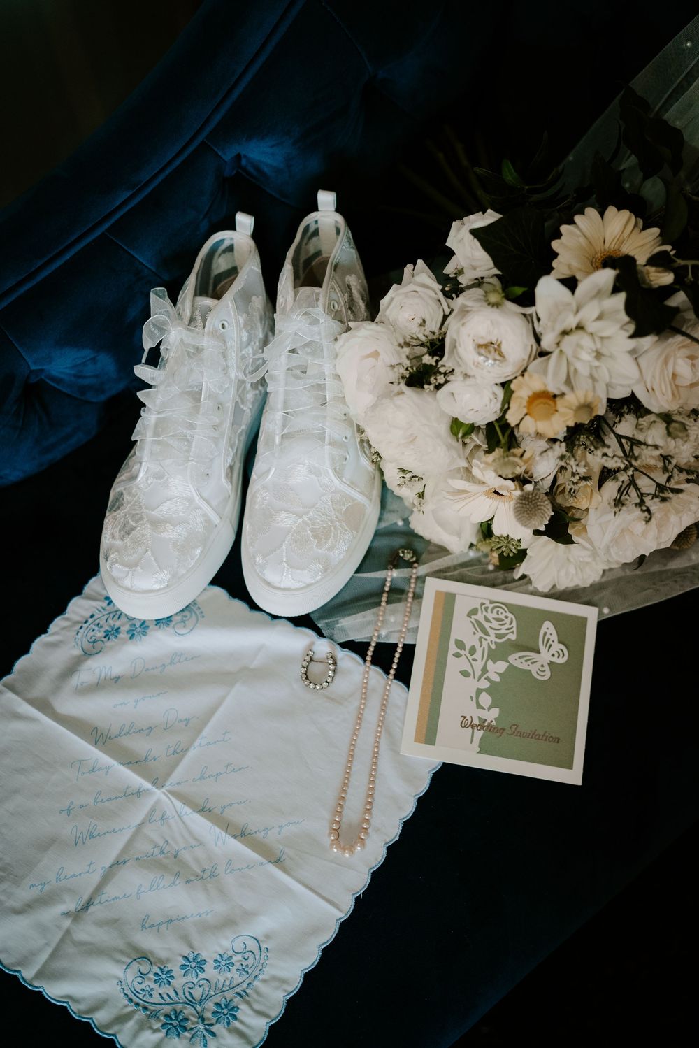 Flat lay of bridal accessories including lace sneakers, pearl necklace, bouquet, and embroidered handkerchief.