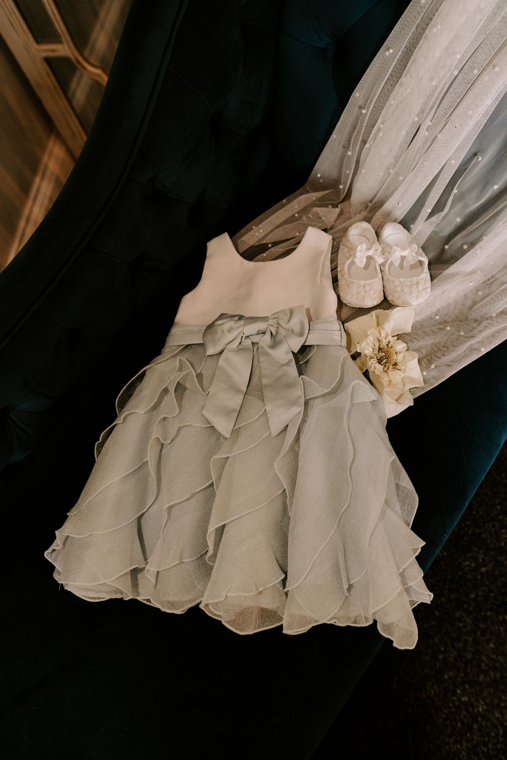 Delicate flower girl dress with sage green ruffled skirt and bow displayed alongside white satin shoes.