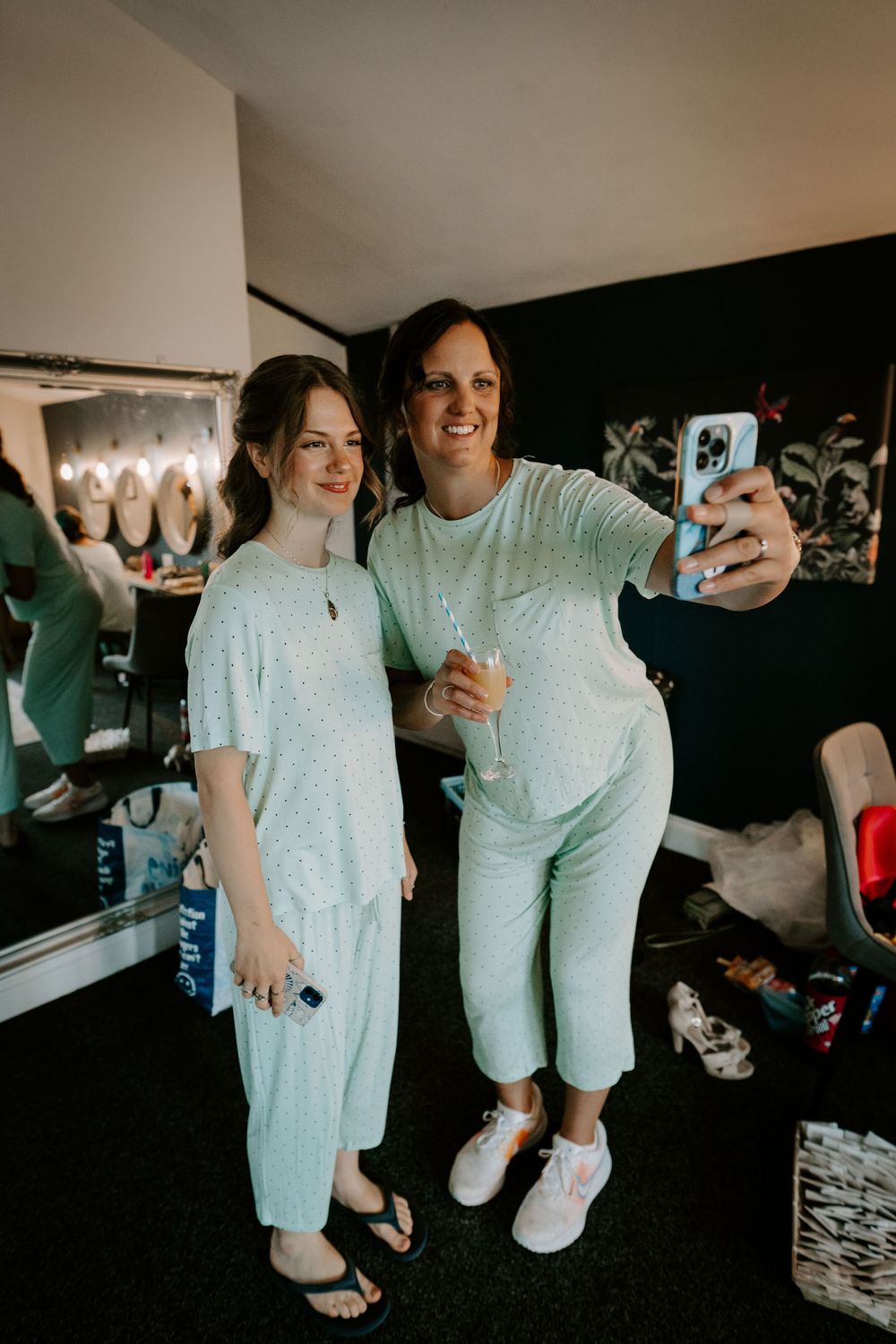 Two bridesmaids in matching mint pajamas taking selfie with champagne in bridal suite.