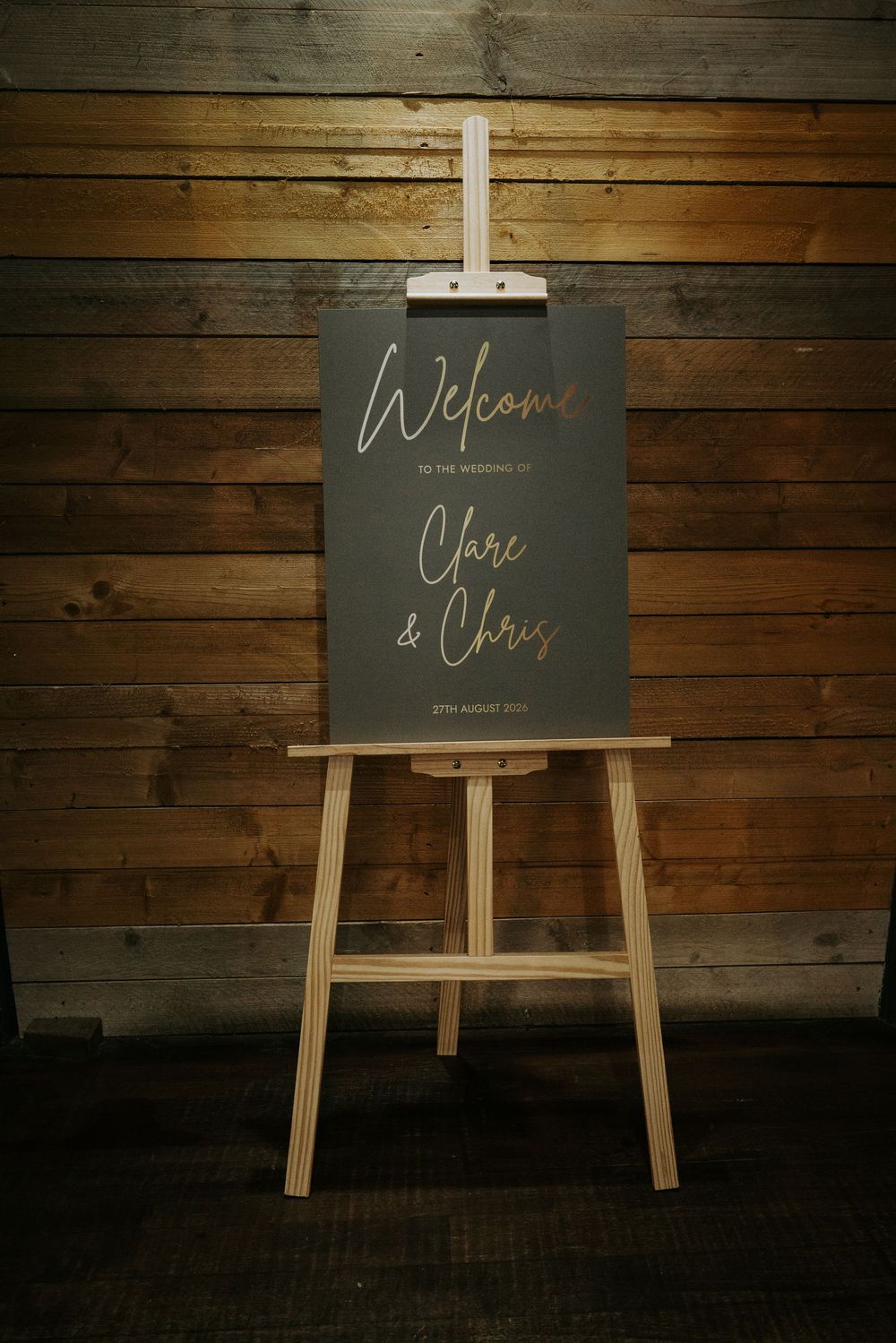 Gold calligraphy welcome sign for Clare and Chris wedding on wooden easel against rustic wall.
