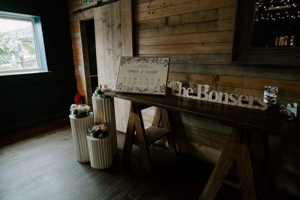 Rustic wooden wedding welcome display with The Bonsers sign and floral arrangements in barn venue.