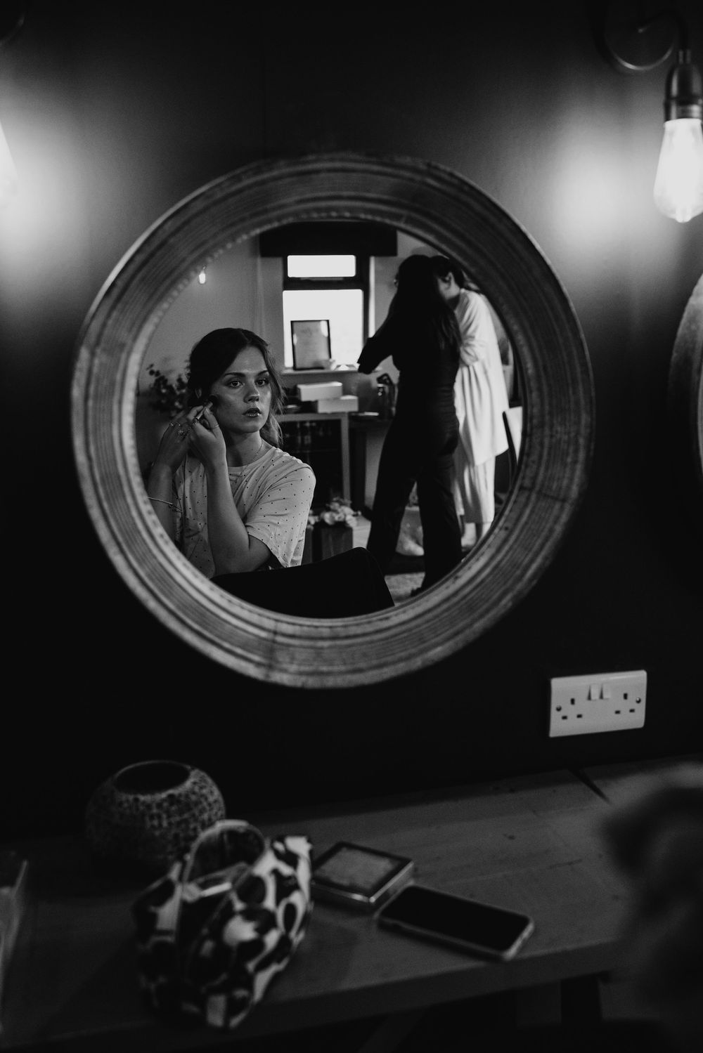 Black and white mirror reflection of bridesmaid adjusting earring with bride in robe in background.
