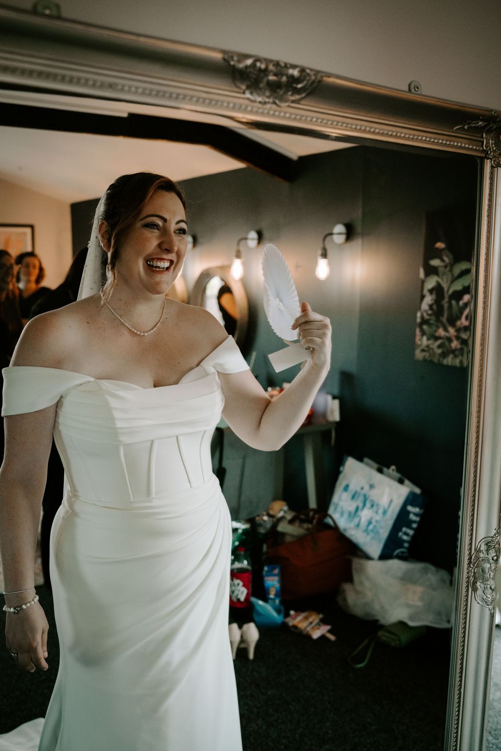 Bride in off-shoulder white gown laughing joyfully while holding a mirror during wedding preparations.
