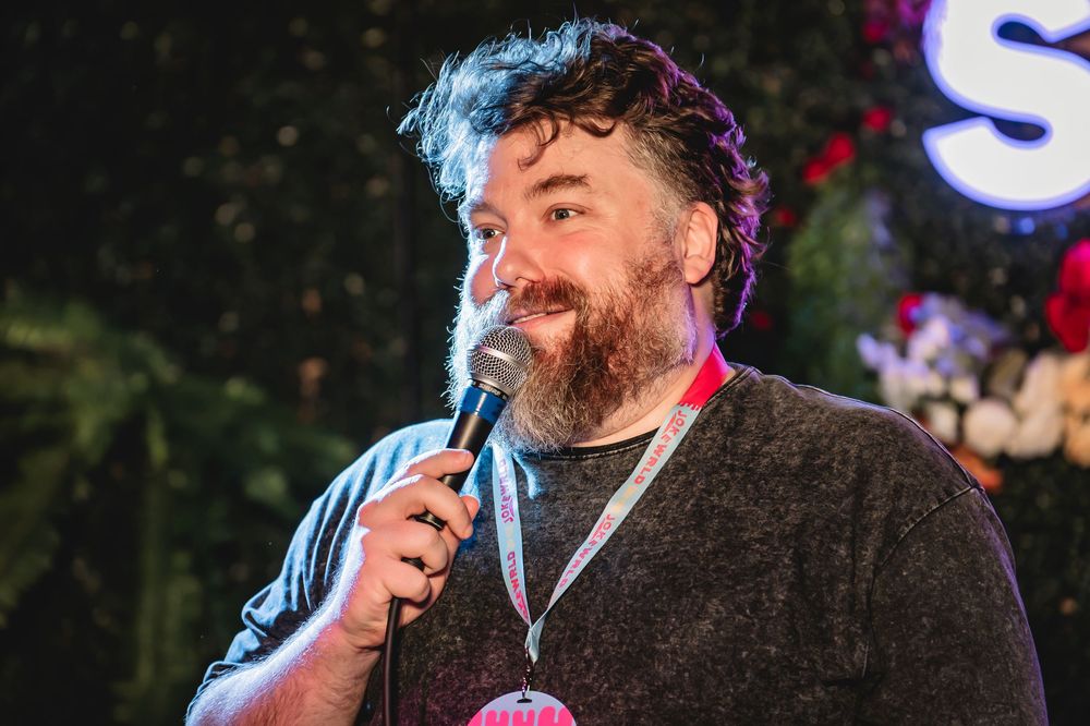 Dan St Germain, bearded man performing stand-up comedy on stage, holding a microphone with colorful lights in background.
