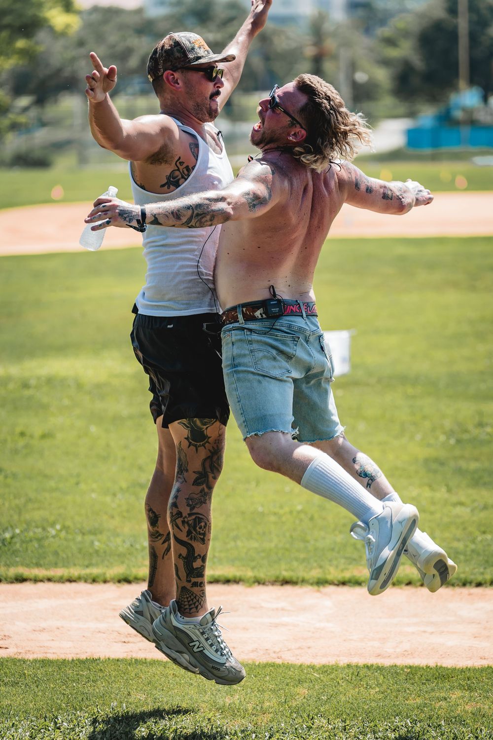 Uncle Lazer. Two tattooed men jumping and celebrating outdoors on a baseball field on a sunny day.
