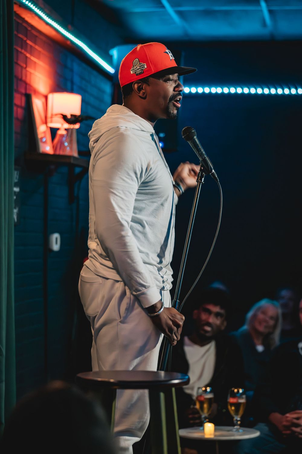 Comedian in white hoodie and red cap performs stand-up at microphone stand before live audience.