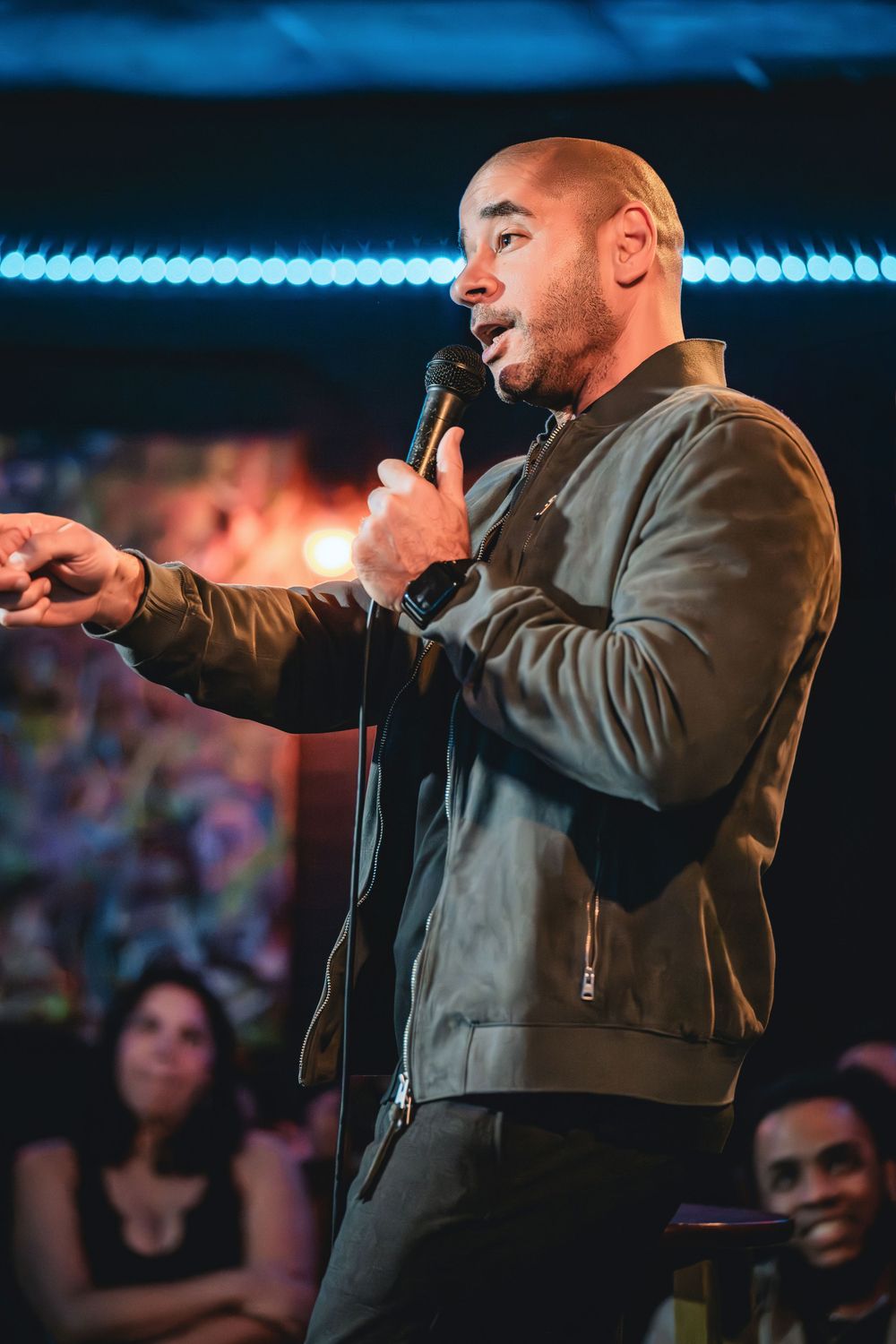 Dino Vigo the comedian. A bald man in an olive jacket performs stand-up comedy on stage, holding a microphone with an audience behind him.
