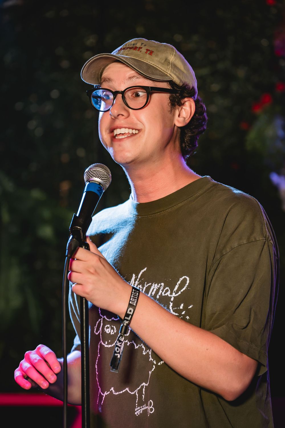 Jack Shaw a young comedian performing stand-up at a microphone, wearing glasses, a cap, and an olive green t-shirt.