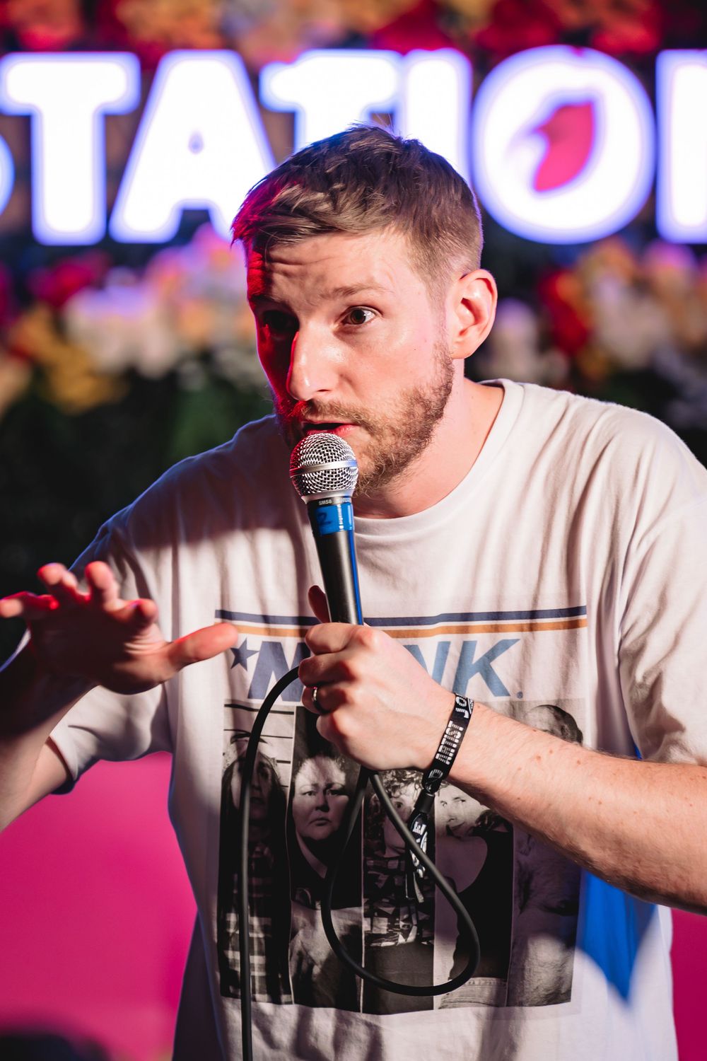Jeremiah Watkins, A bearded man in a graphic t-shirt holds a microphone on stage at a comedy show with colorful lights behind him.