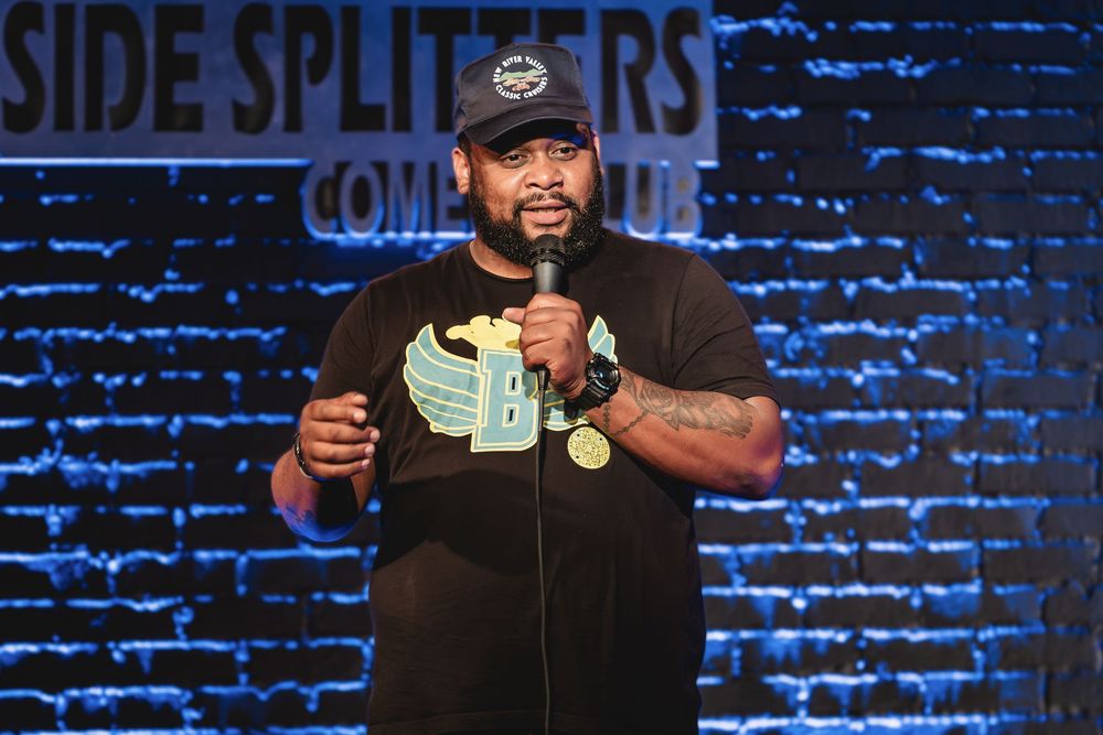 Jordan B the Comedian performing stand-up on stage at Side Splitters Comedy Club, holding a microphone under blue brick wall lighting.