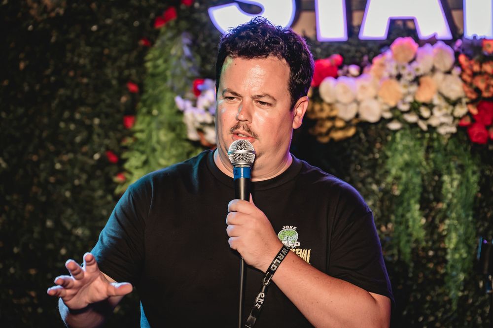 Michael Turner the comedian performing stand-up on stage, holding microphone, wearing black t-shirt, with floral backdrop at comedy club.