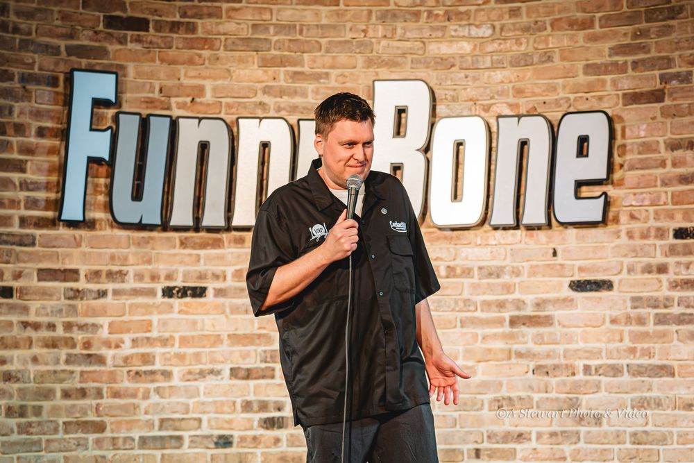 Comedian performing stand-up on stage at Funny Bone comedy club, holding microphone in front of brick wall.