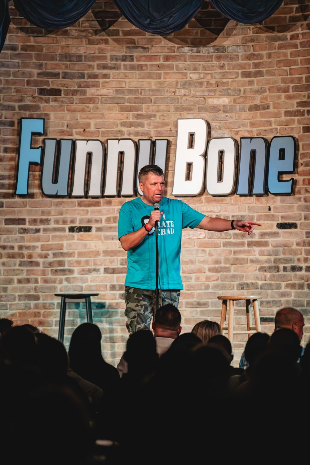 Comedian performing stand-up on stage at Funny Bone comedy club, holding microphone in teal shirt.