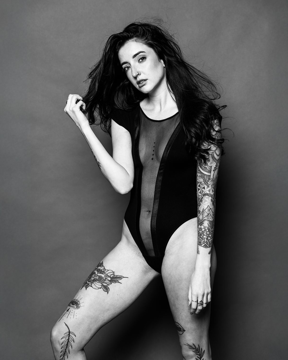 Black and white portrait of a tattooed woman in a black bodysuit posing confidently in a studio.