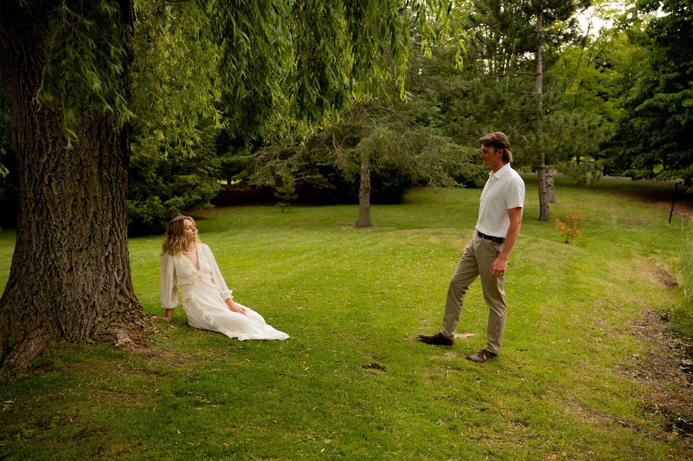 A woman in a white dress sits under a large tree while a man in casual attire stands nearby on a lush green lawn.