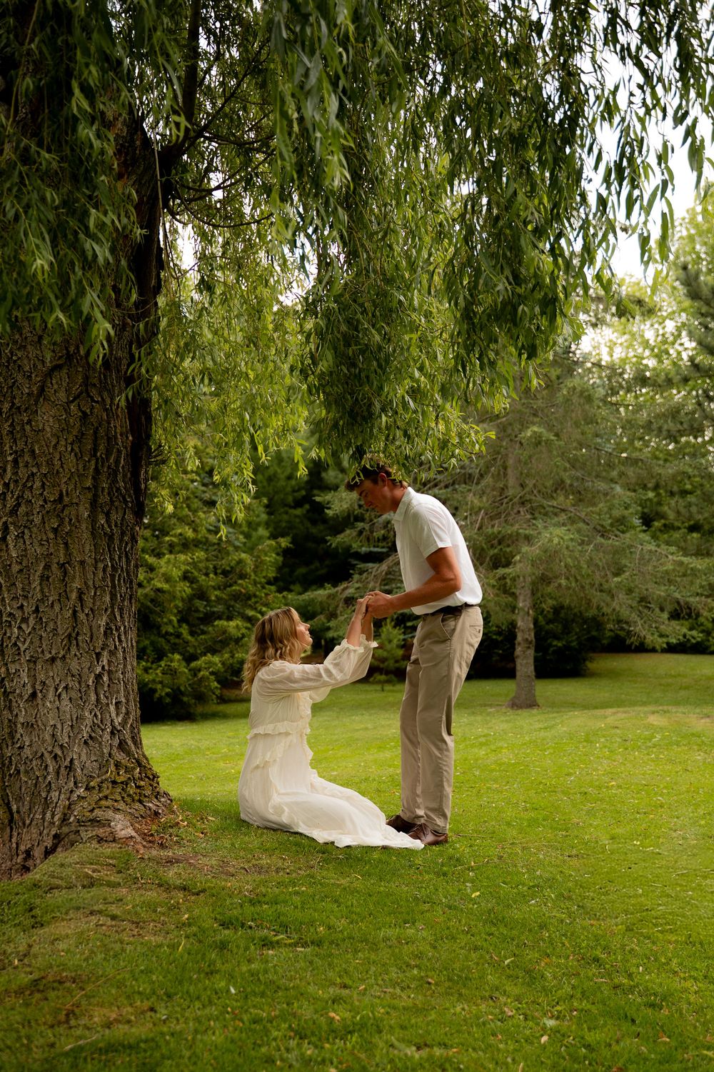 Man helps woman in white dress rise from grass under weeping willow tree in lush green park.