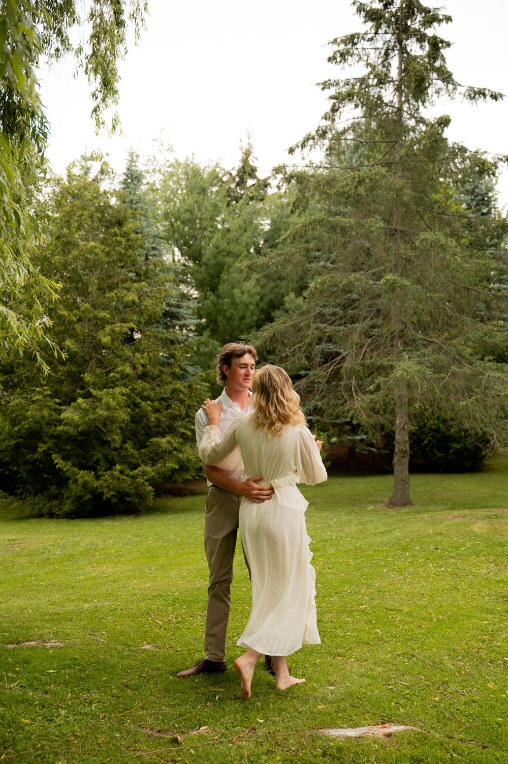 A couple dances together on green grass surrounded by tall evergreen trees in a lush garden setting.
