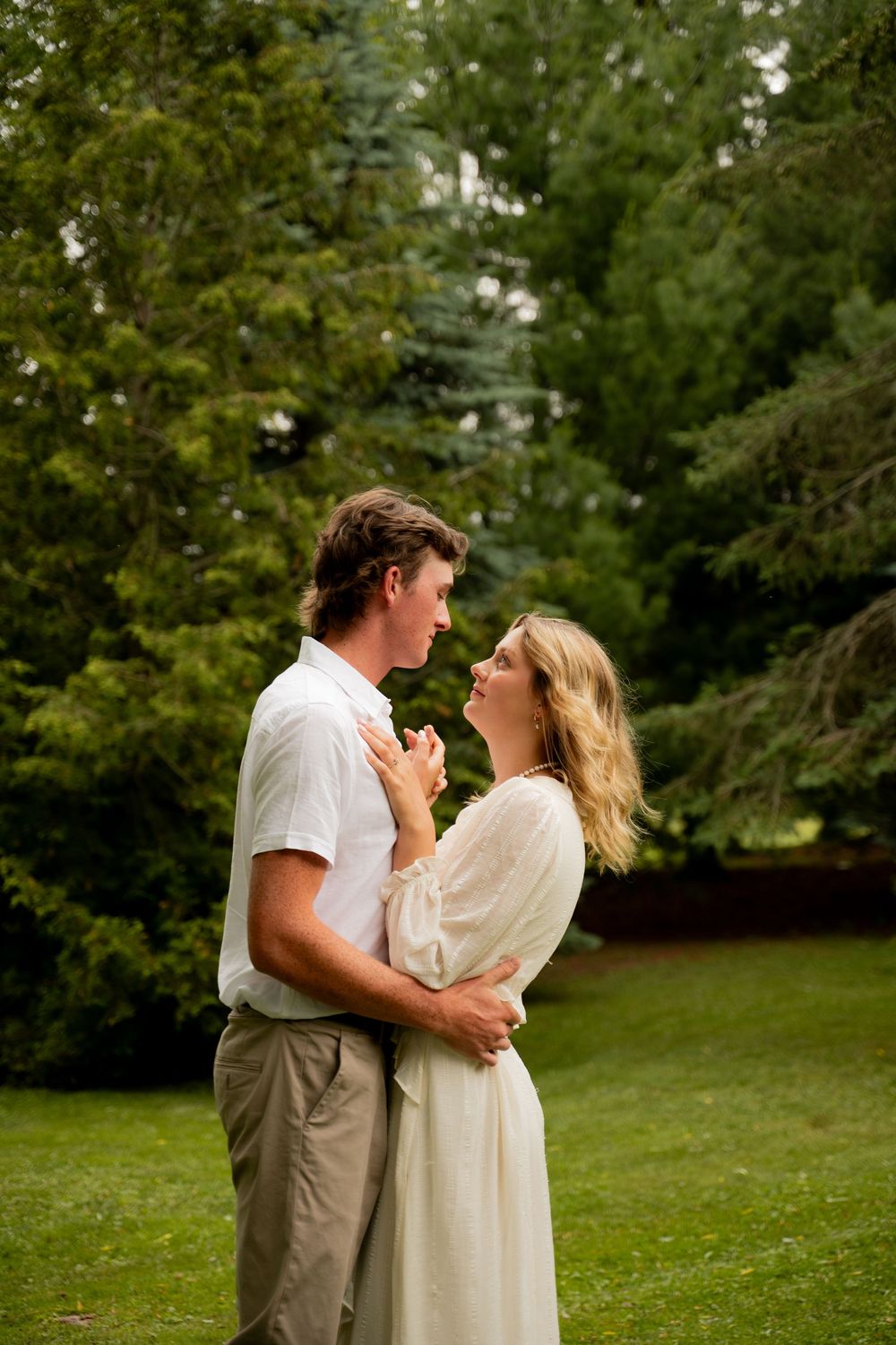 Couple embracing in lush green garden, woman in white dress and man in white shirt sharing romantic moment outdoors.