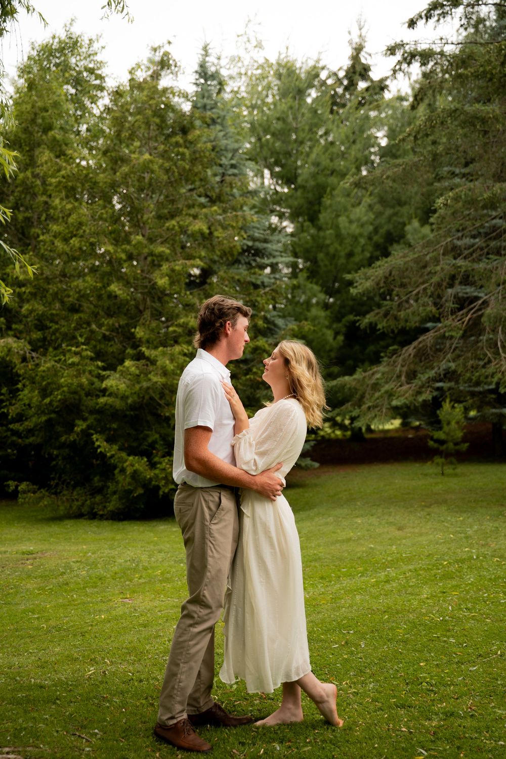 A couple embraces on a lush green lawn surrounded by tall trees in a romantic outdoor setting.