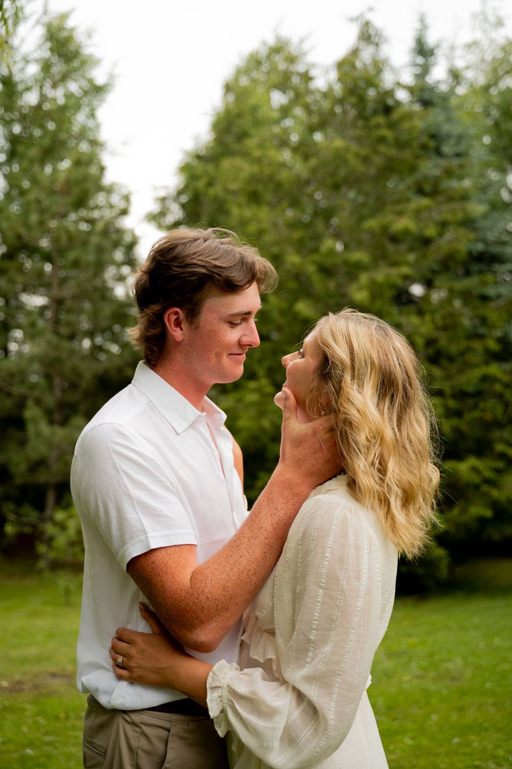 A young couple shares a romantic moment outdoors, embracing near lush green trees on a cloudy day.