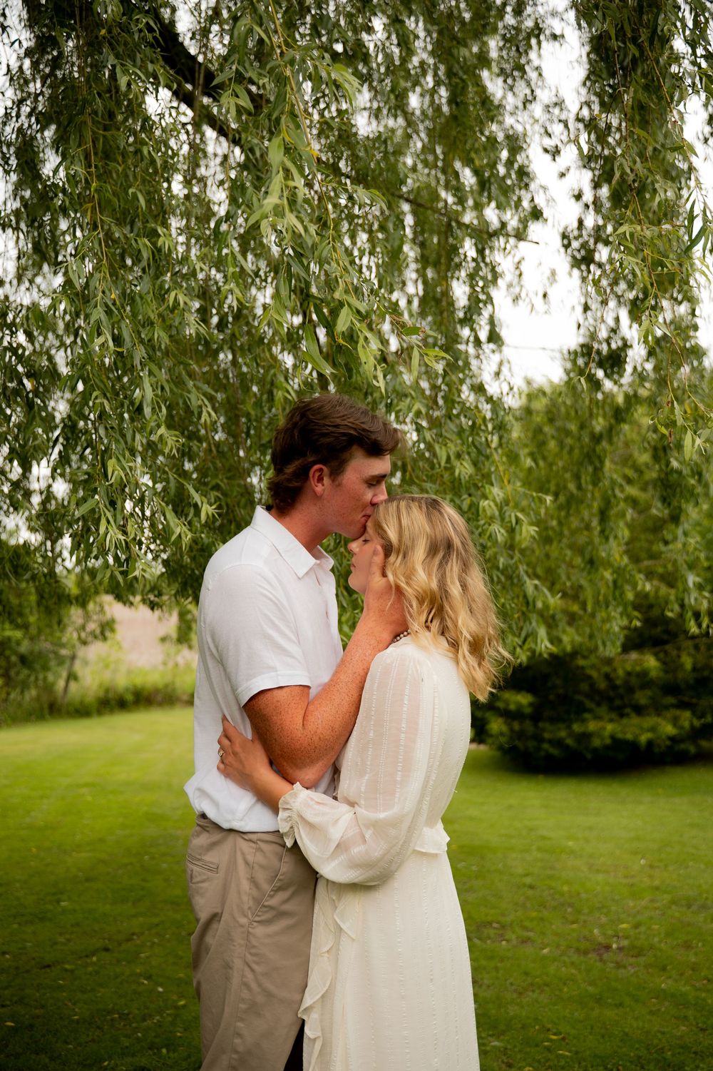 Couple sharing a romantic kiss under a weeping willow tree in a lush green garden, dressed in white and khaki.