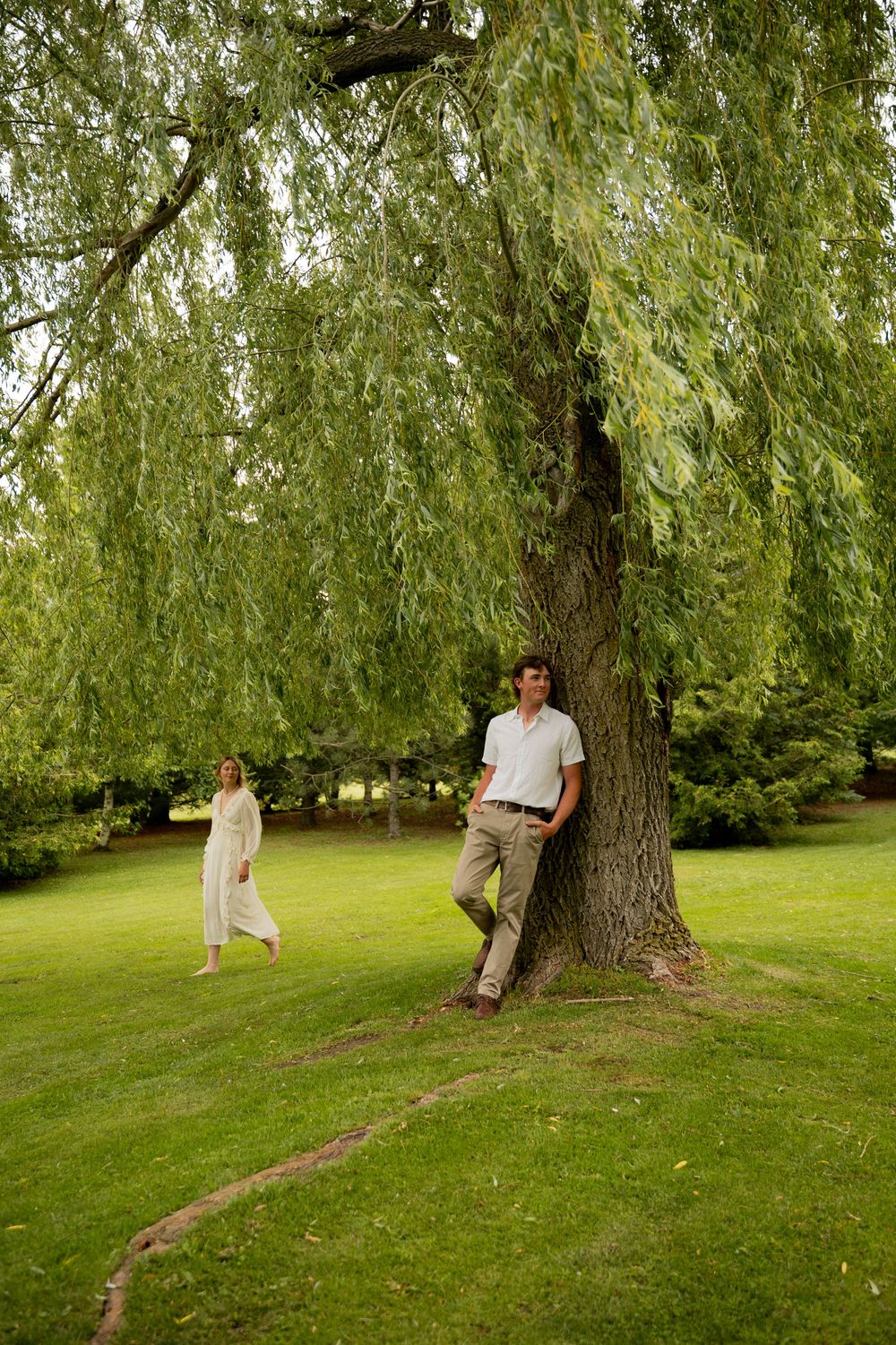 Man leaning against a weeping willow tree in a lush green park with a woman walking in the background.