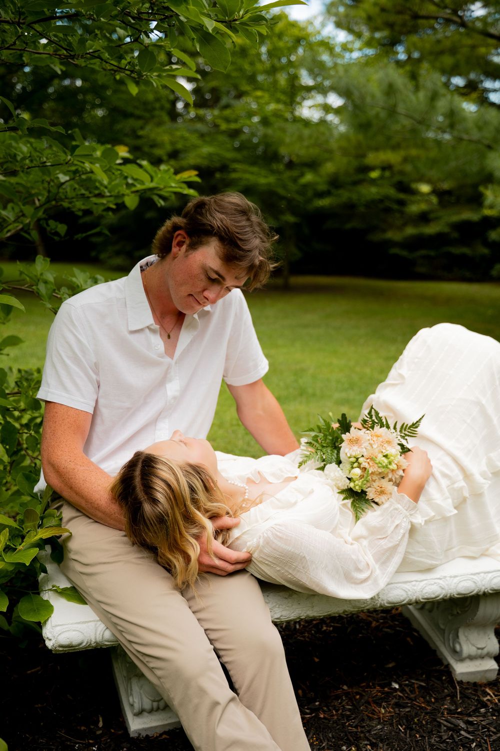 Bride in white dress lying on bench with bouquet, groom in white shirt gazing at her in lush garden.