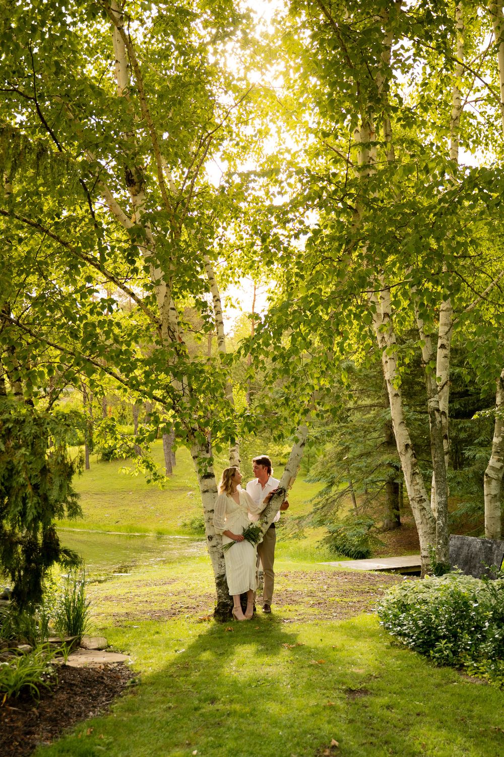 A couple in wedding attire stands romantically among birch trees in a lush green garden setting.