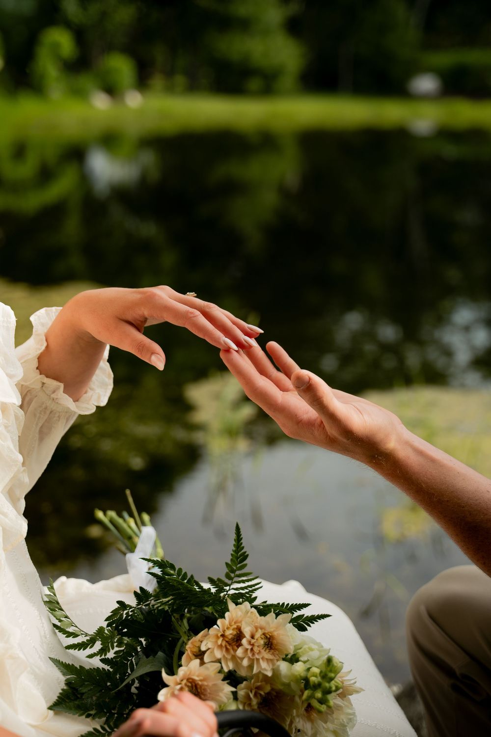 Bride and groom gently touching hands near a serene pond, with a bouquet of ferns and flowers in the foreground.