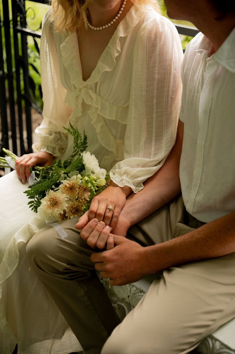 Bride and groom holding hands showing wedding rings, bride holding white floral bouquet in elegant sheer dress.