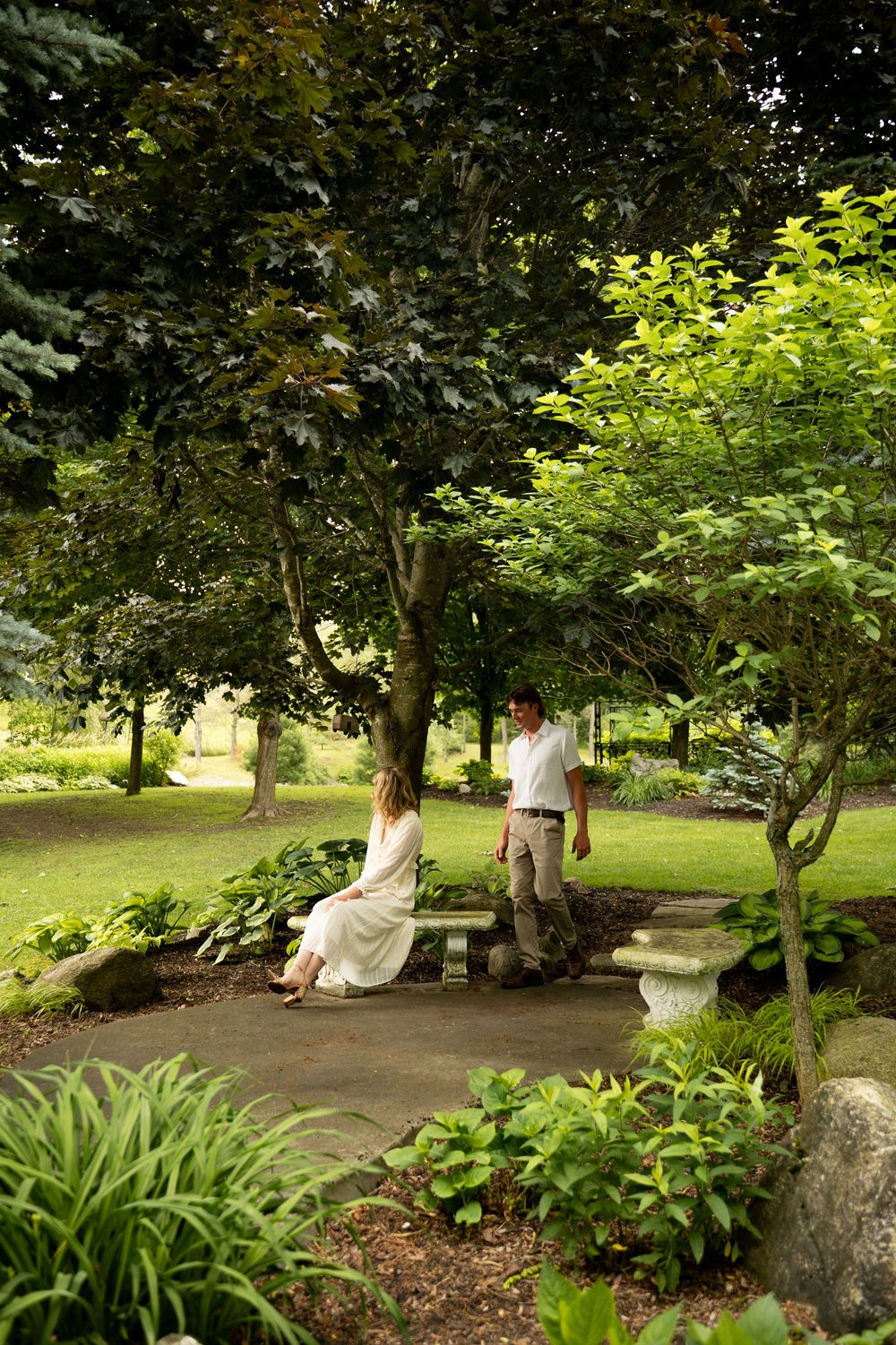 Couple in lush garden, woman in white dress seated on bench while man stands nearby among green trees.