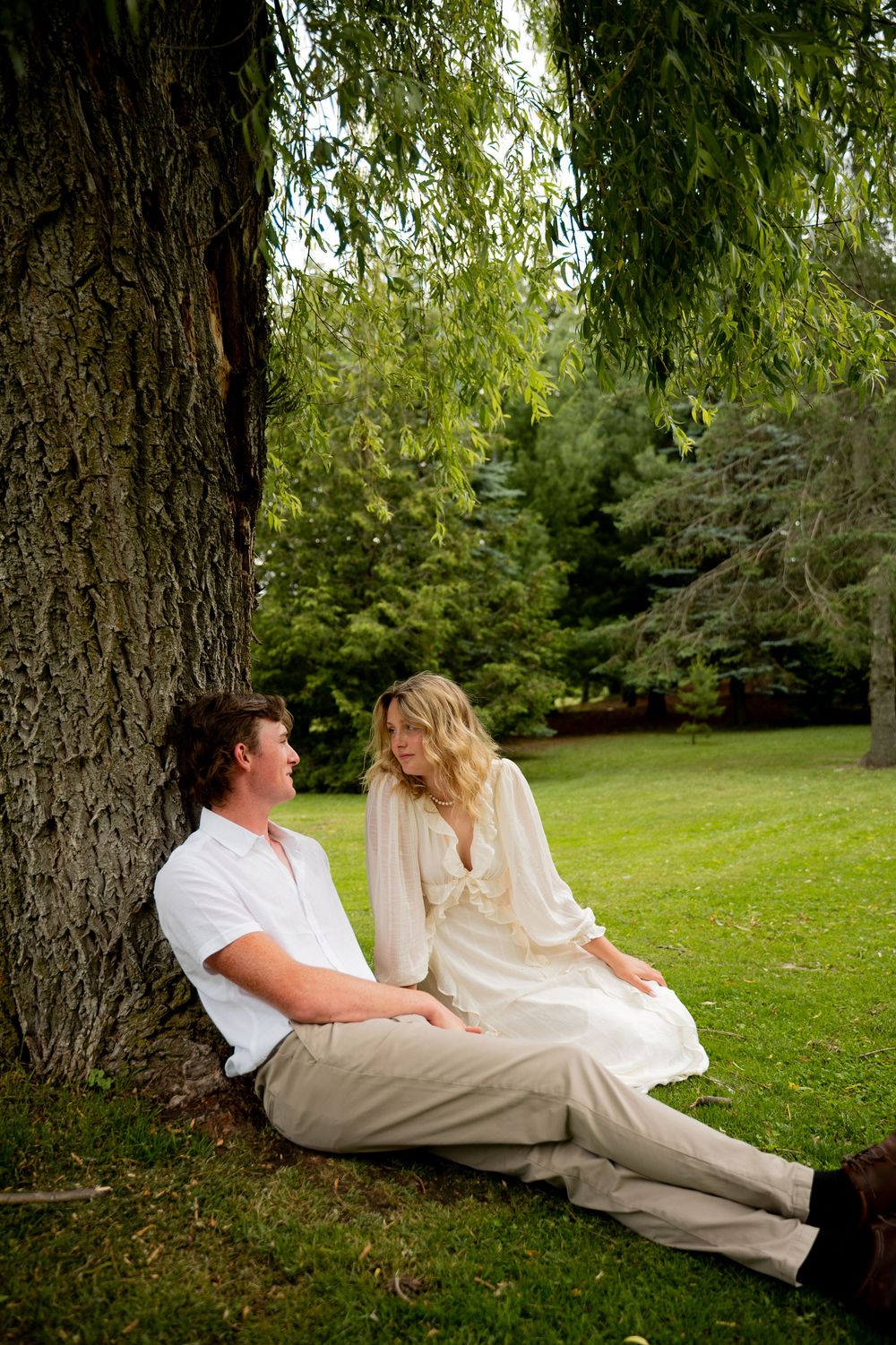 Couple sitting together under a large willow tree on green grass in a lush park setting.