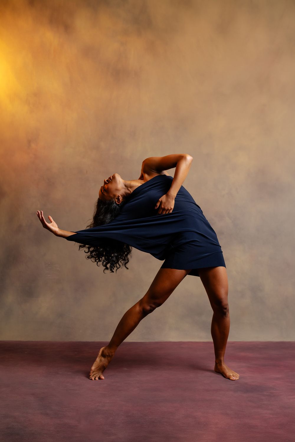 A dancer in a navy blue dress strikes a dramatic backbend pose in a warmly lit studio.