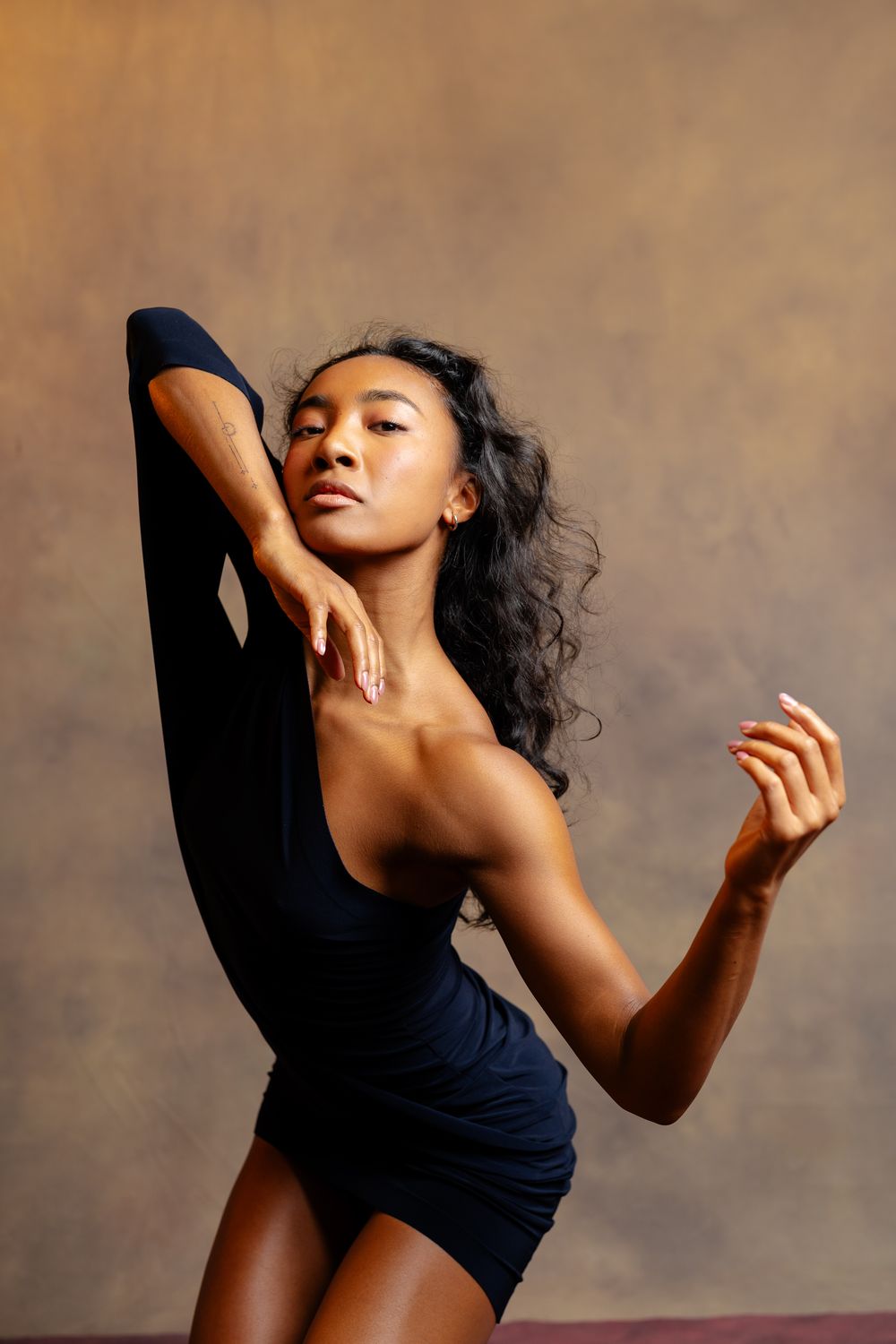 A graceful Black female dancer in a black leotard poses elegantly with leg raised, against a warm studio backdrop.