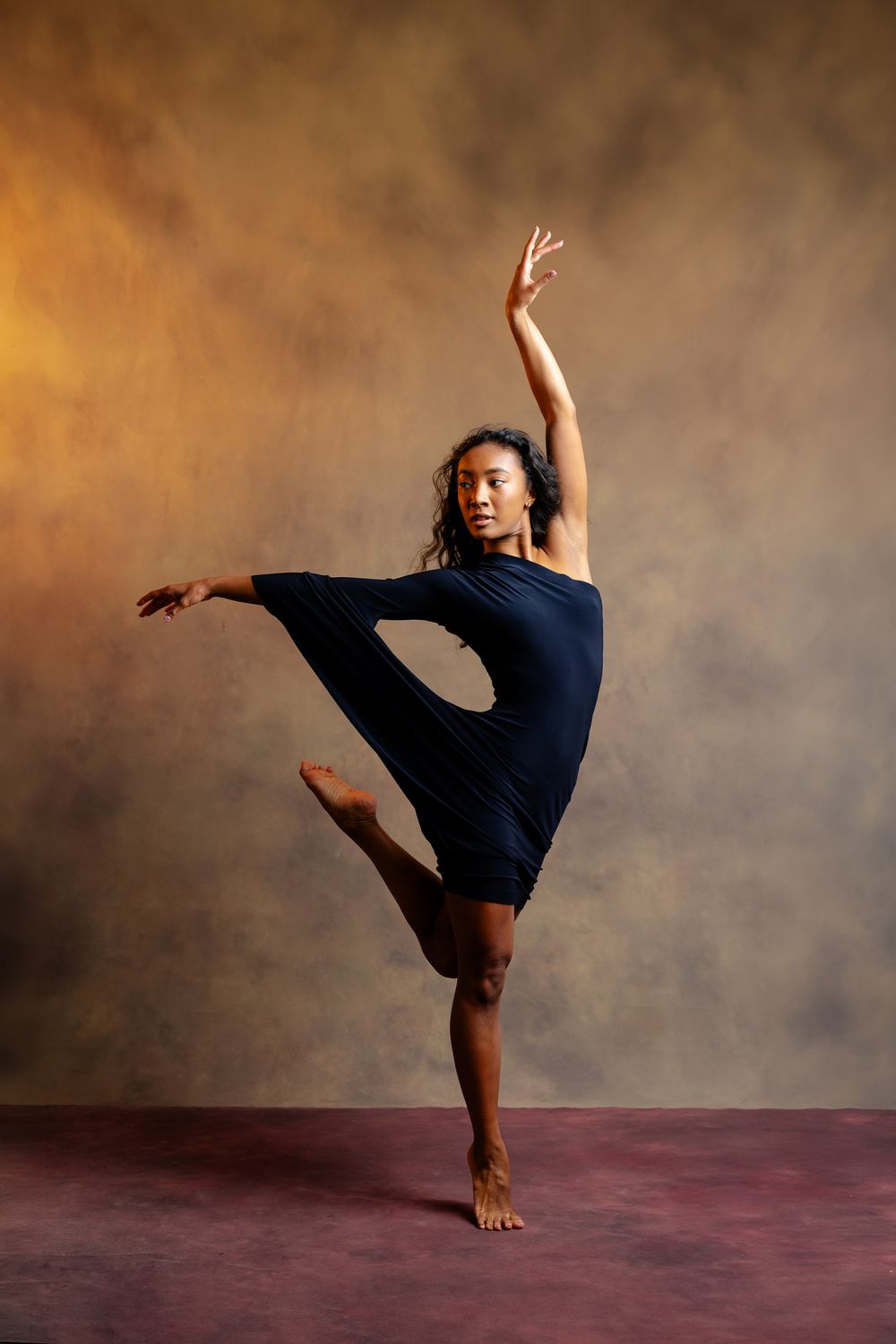 A graceful dancer in a black dress balances on one leg with arms raised in an elegant studio pose.