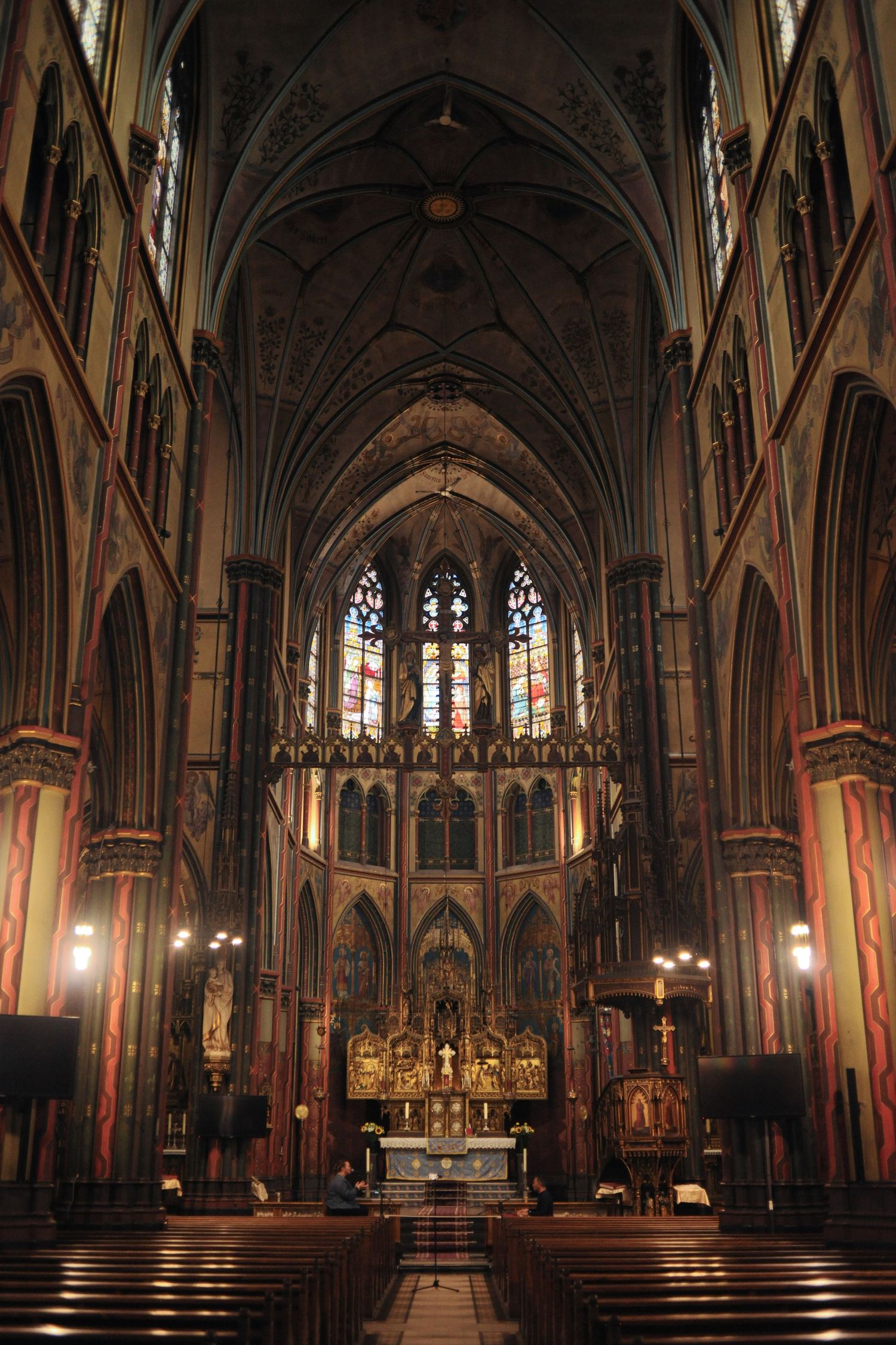 Gothic cathedral interior with ornate altar, stained glass windows, vaulted ceilings, and wooden pews in dim lighting.