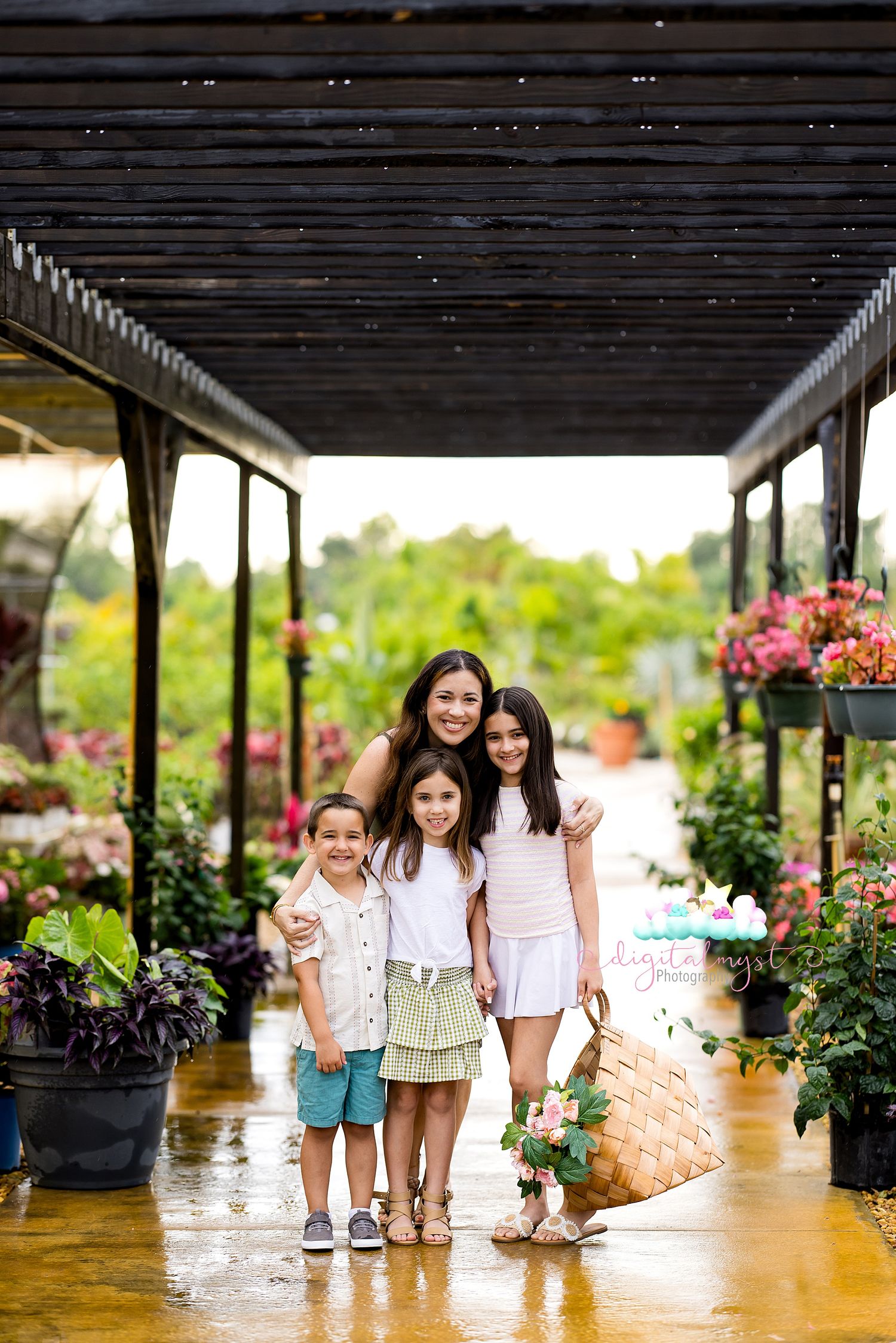 A mother and two children smile together under a pergola at a lush flower nursery after rain.