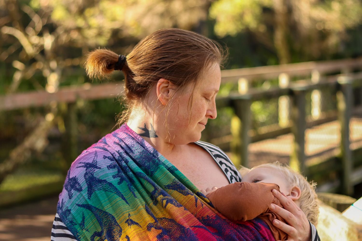 Smiling mother with tattoo holding a sleeping blonde toddler in a floral baby carrier outdoors.