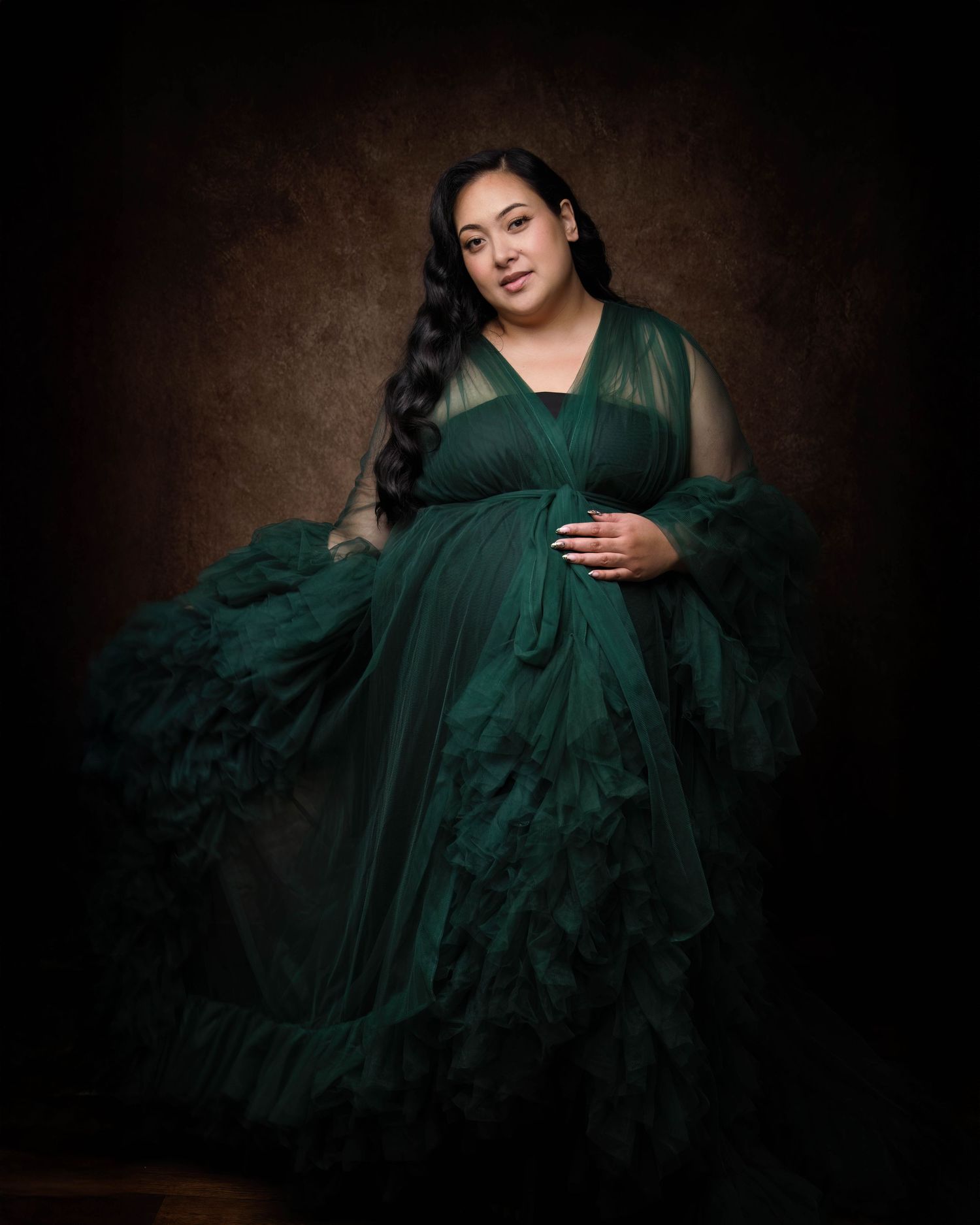 Pregnant woman in elegant dark green tulle maternity gown poses gracefully against a moody studio backdrop.