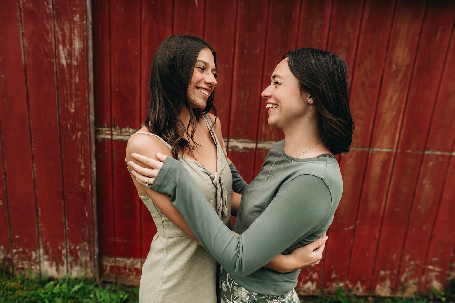 Two smiling women embracing each other outdoors in front of a rustic red barn wall.