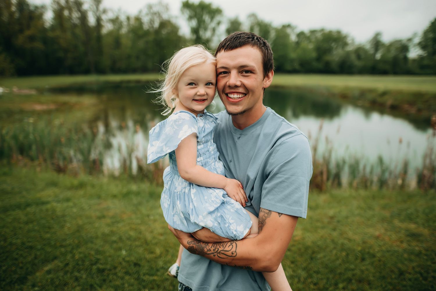 A smiling young man holds a happy toddler girl in a blue dress near a serene pond surrounded by lush greenery.