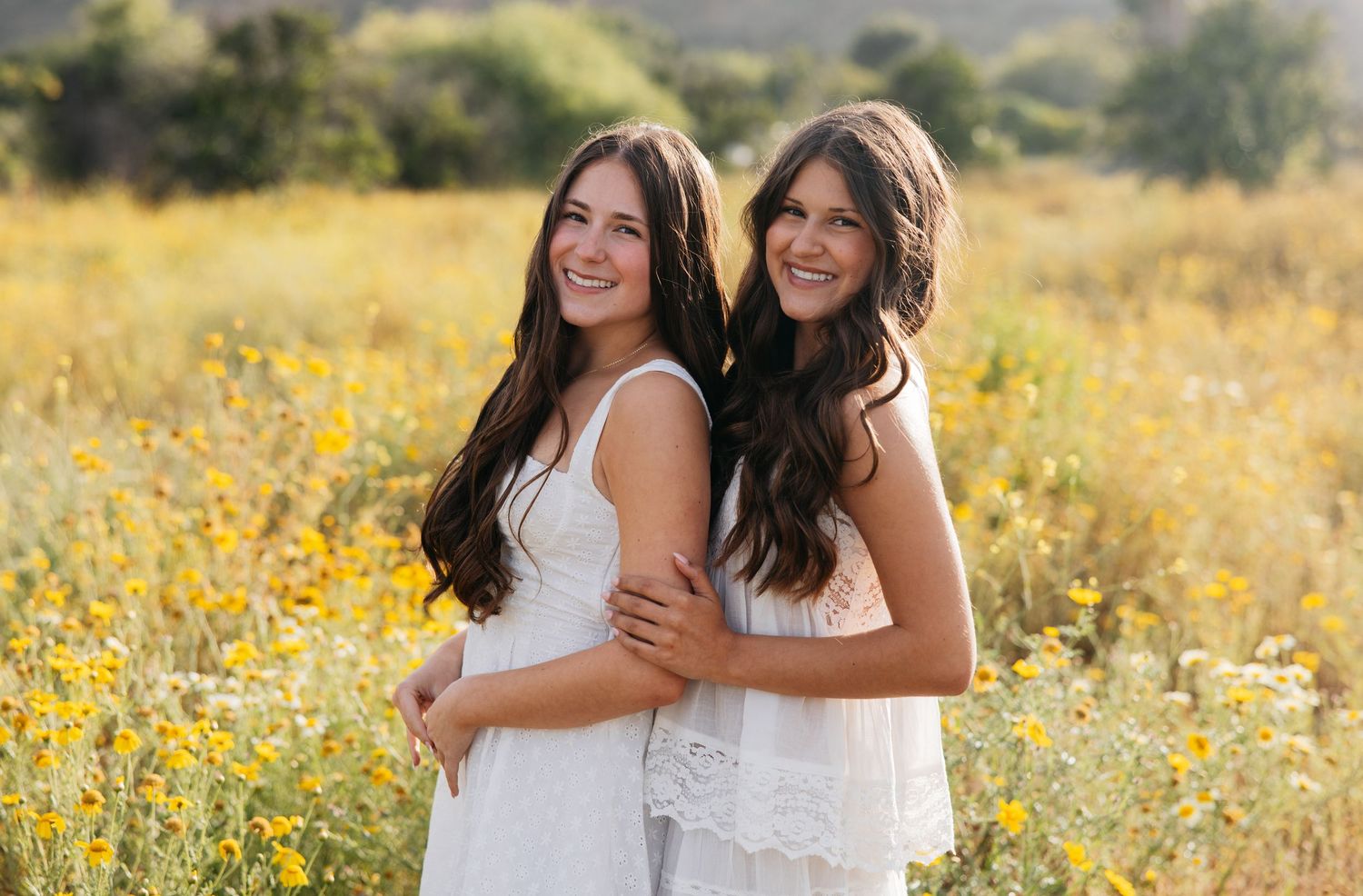 Two young women in white dresses smiling together in a sunny wildflower field.