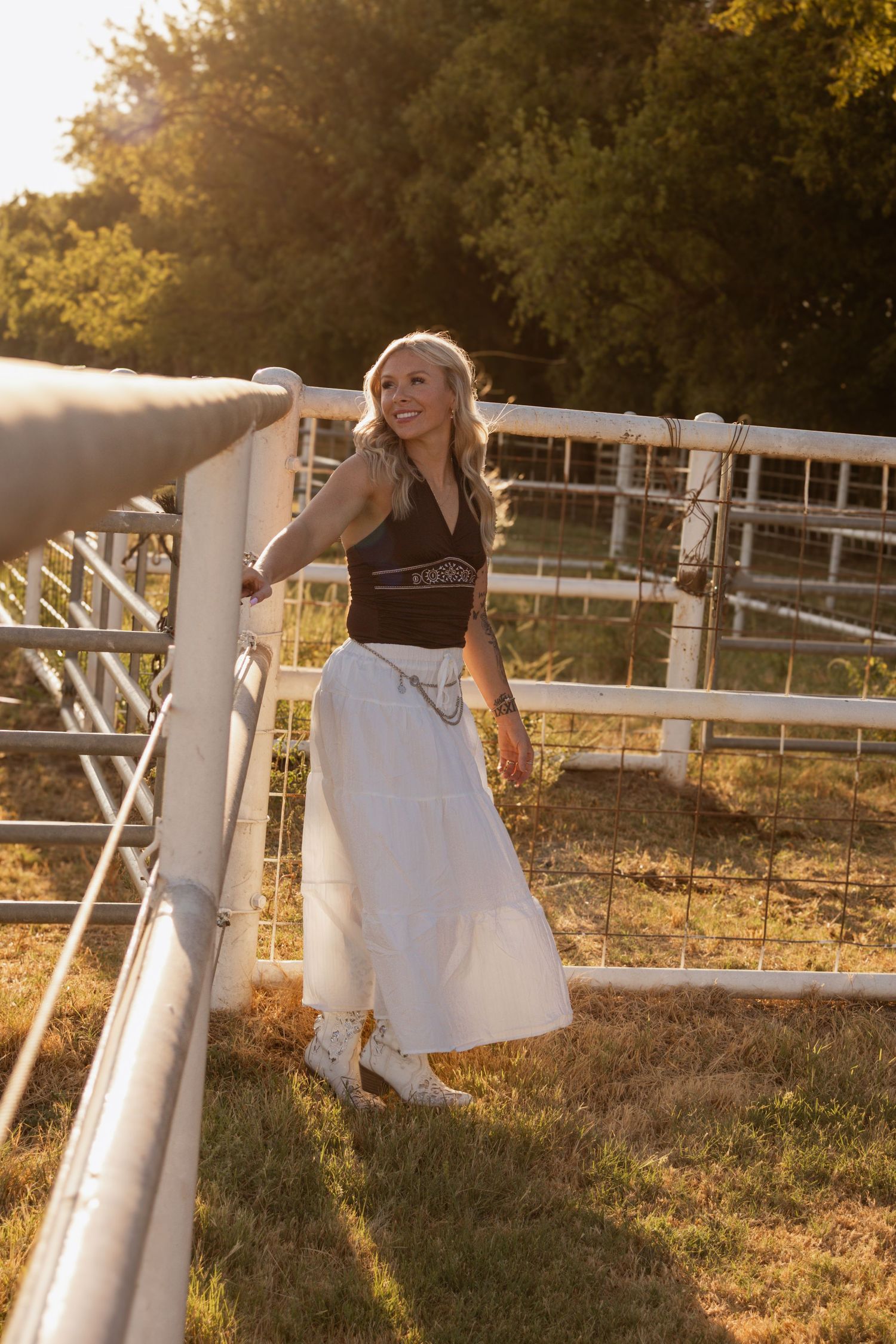 Woman in a flowing white dress posing by a fence during a warm golden hour portrait session.