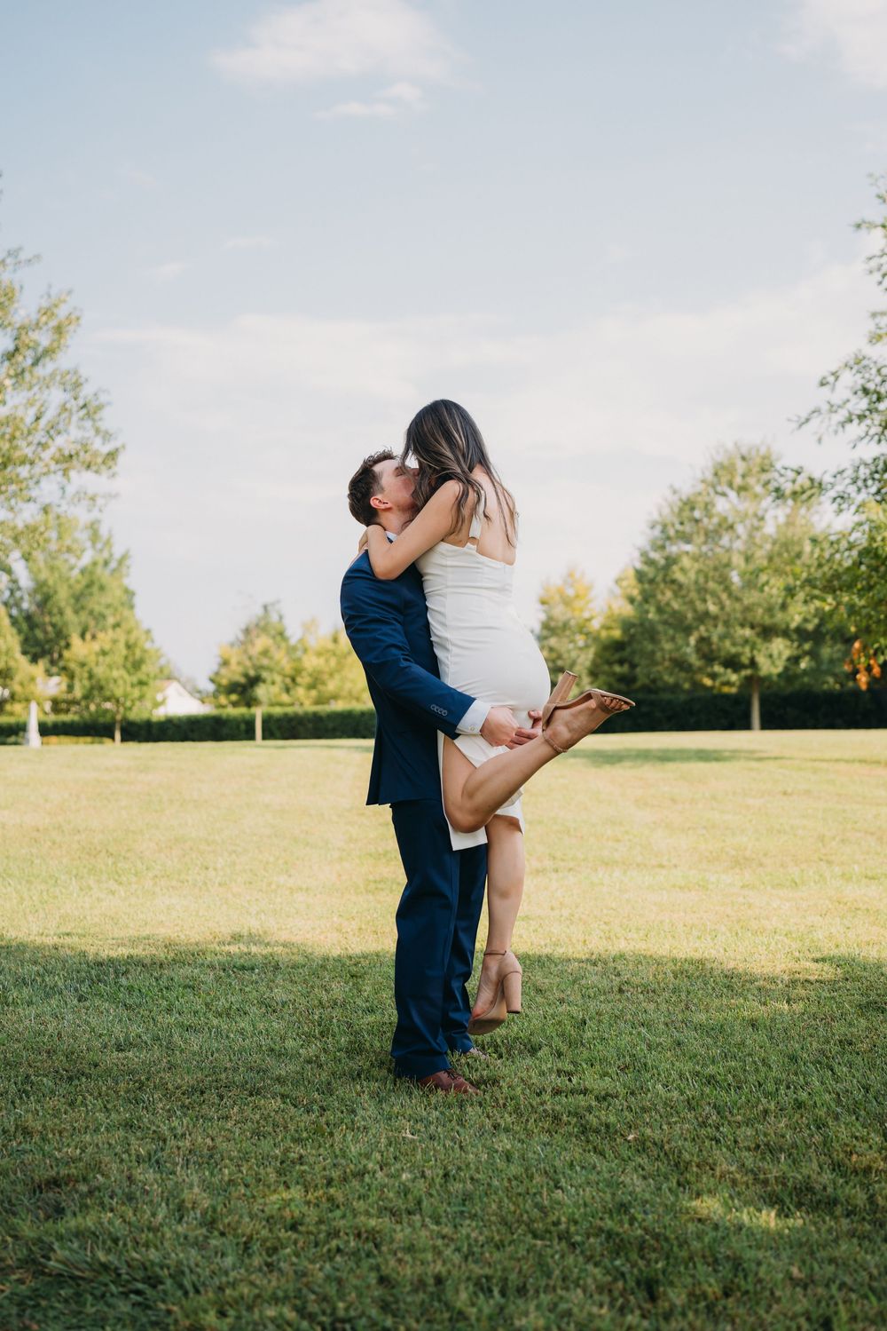 Man in navy suit lifts woman in white dress for a romantic kiss on a lush green lawn outdoors.