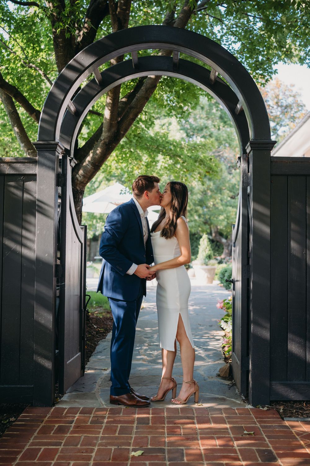 Couple sharing a romantic kiss under a black arched garden gate, man in blue suit, woman in white dress.