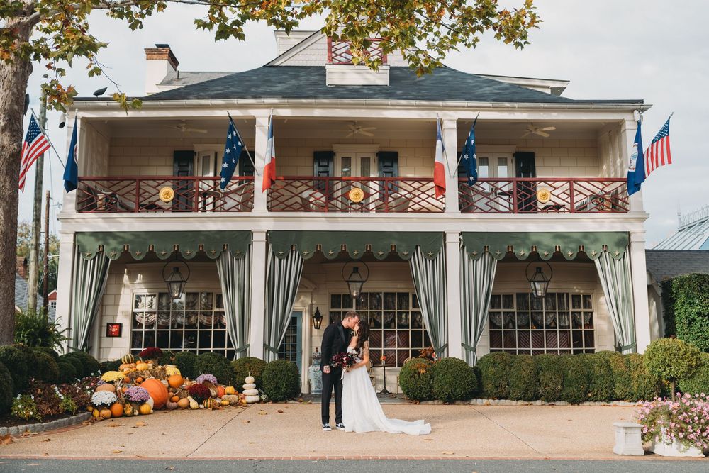 Bride and groom kissing outside a charming two-story venue decorated with fall pumpkins and flags on a sunny autumn day.