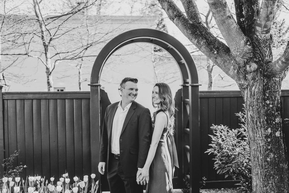 Black and white photo of a smiling couple standing under a garden arch with tulips and bare trees.