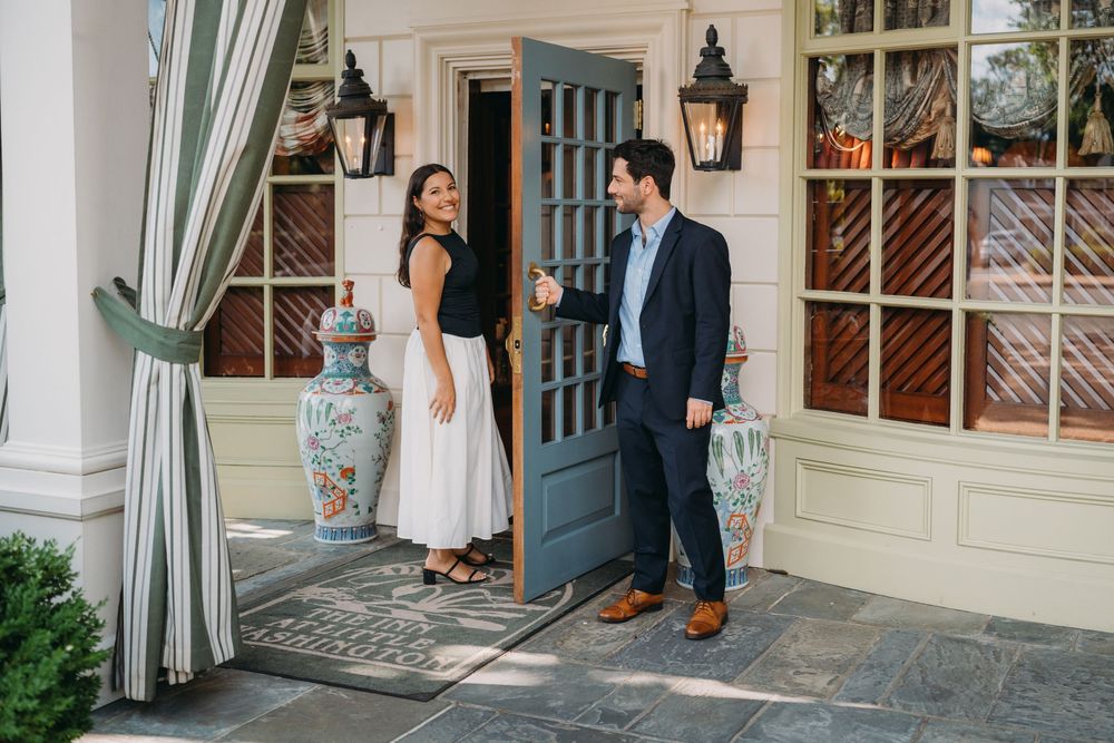 Couple standing at elegant blue French door entrance of upscale home, man in navy suit, woman in white dress.